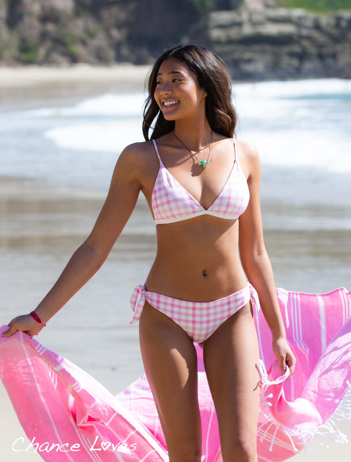 A smiling 19 year old girl walking on the beach in Laguna three arch bay wearing a gingham style pink and white swimsuit by Chance Loves