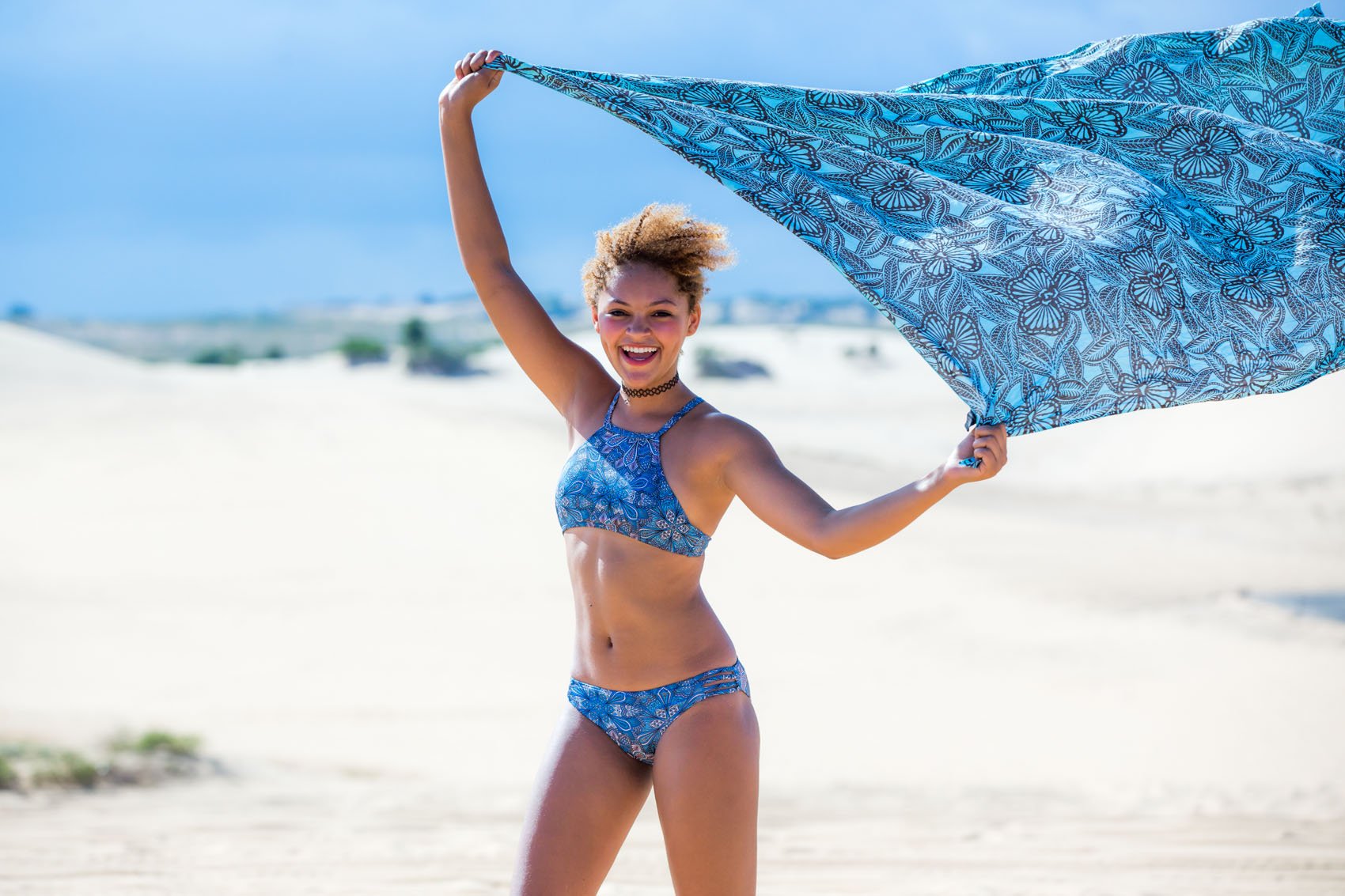 Teenage girl laughing while holding up a scarf on a white sandy beach in a blue swimsuit