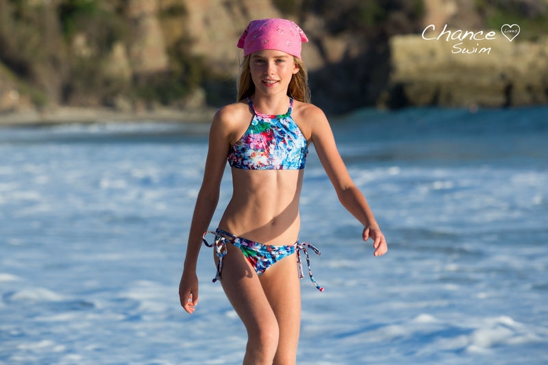 A 10 year old girl walking on the beach wearing a pink bandana in her hair, and a colorful matching 2-piece bathing-suit